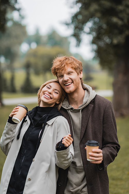 cheerful man and pleased young woman holding coffee to go while laughing in park