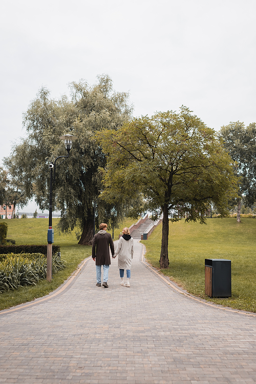 back view of happy redhead man and pleased young woman holding hands while walking in park