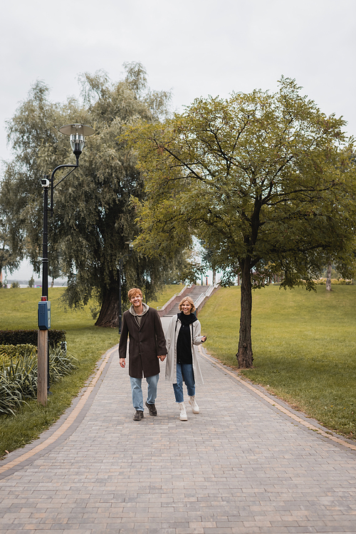 full length of joyful redhead man and pleased young woman in coat holding hands while walking in park