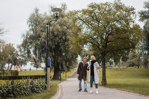 full length of happy redhead man and blonde young woman in coat holding hands while walking in park