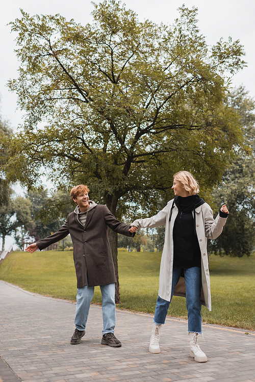 full length of happy man and pleased woman in coats holding hands while walking in park