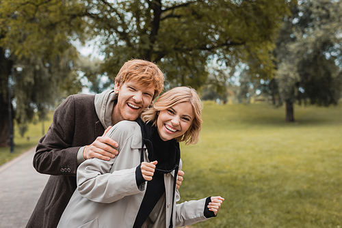 happy man hugging joyful young woman while looking at camera in park