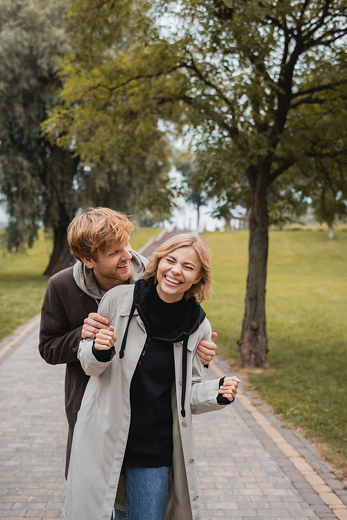 happy man hugging joyful young woman with closed eyes laughing in park