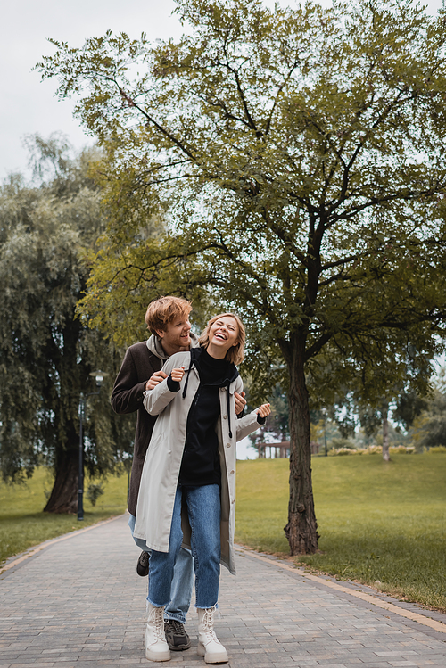 happy man in coat hugging cheerful young woman with closed eyes laughing in park