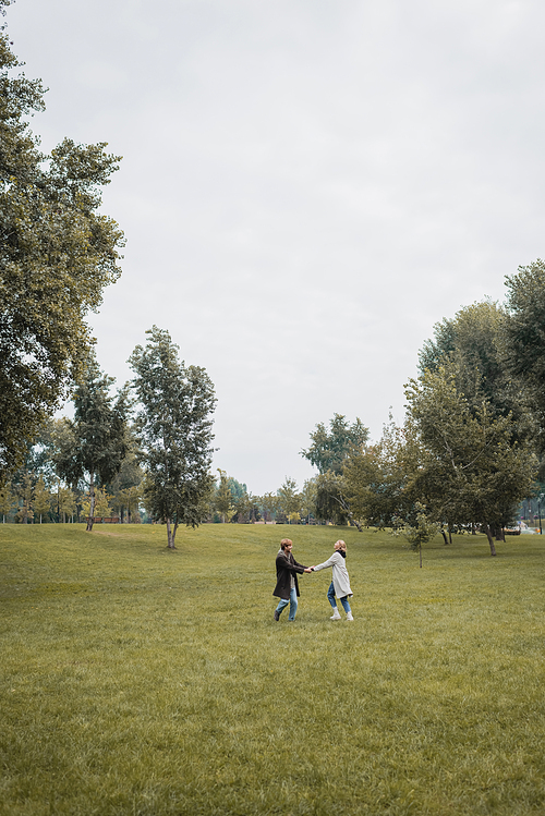 full length of happy man and pleased woman in coats holding hands while spending time in park