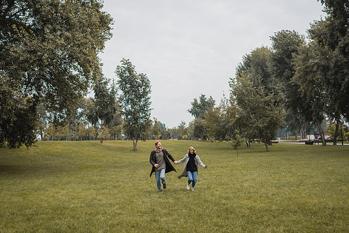 full length of happy man and pleased woman in coats holding hands while running on grass