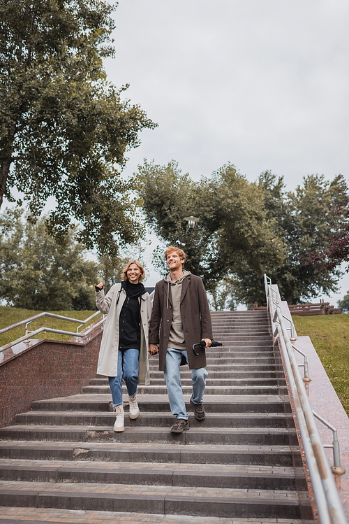 cheerful blonde woman and redhead man with umbrella holding hands while descending stairs in park