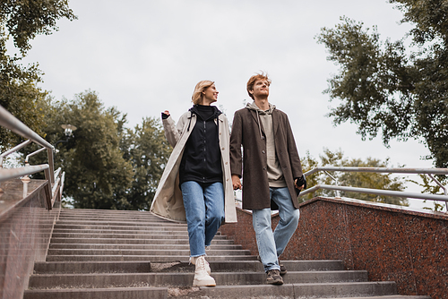 low angle view of cheerful woman and redhead man with umbrella holding hands while descending stairs near park