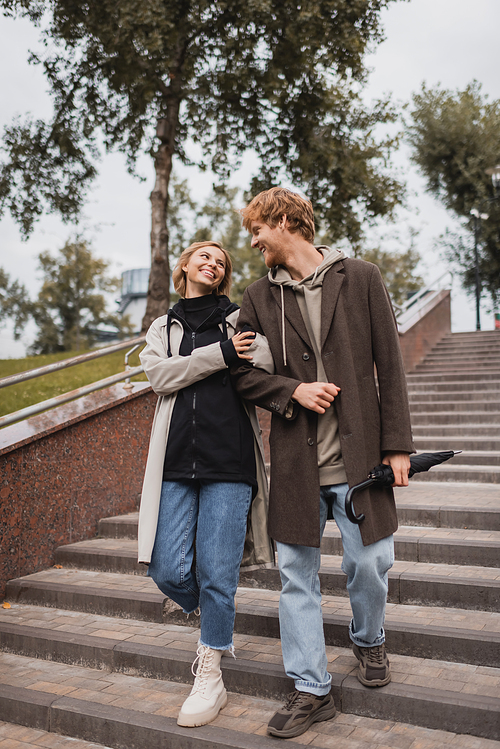 pleased woman and redhead man with umbrella descending stairs in park