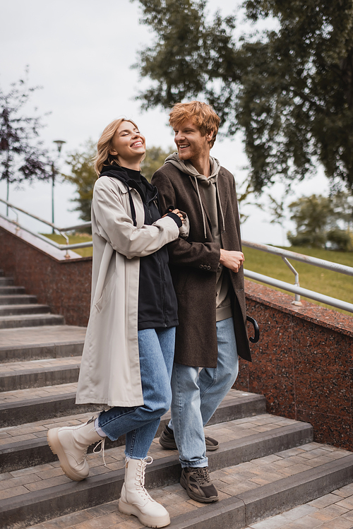 joyful woman and happy redhead man with umbrella descending stairs in park