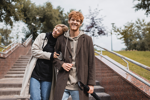 pleased woman leaning on shoulder of redhead man with umbrella while descending stairs in park