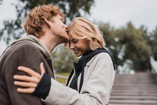 redhead man kissing forehead of happy girlfriend in park