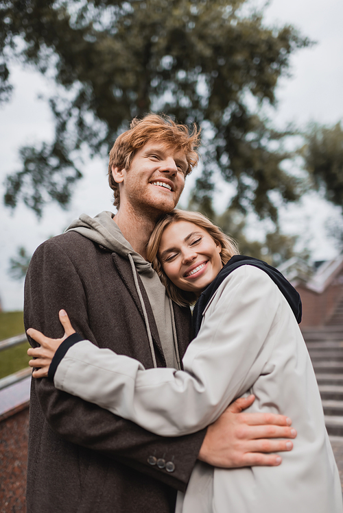 happy young woman hugging redhead boyfriend in autumnal coat in park