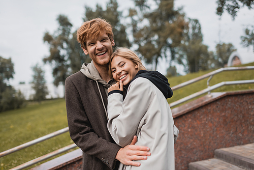 happy young woman hugging with joyful redhead boyfriend in autumnal coat in park