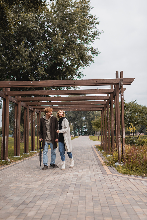 full length of happy young couple in autumnal coats walking under multiple arch in park