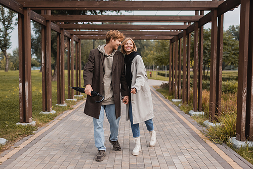 full length of happy young couple in autumnal coats holding hands while walking under multiple arch in park