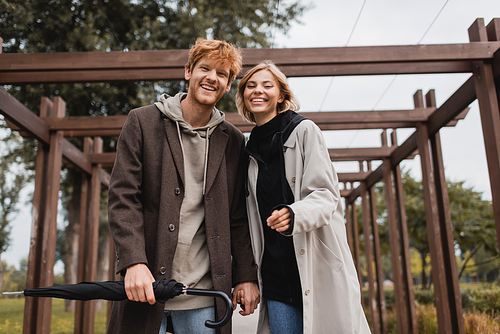 cheerful young couple in autumnal coats holding hands while walking under multiple arch in park