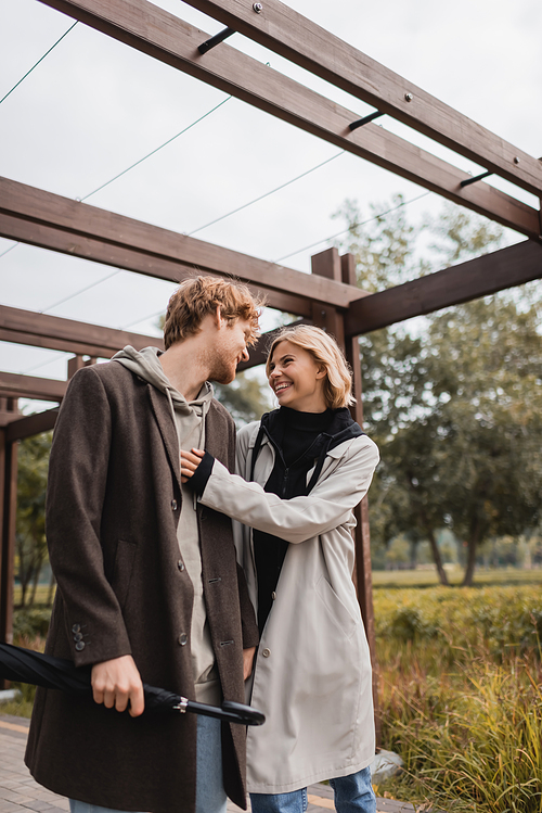 positive young couple in autumnal coats laughing while walking under arch in park