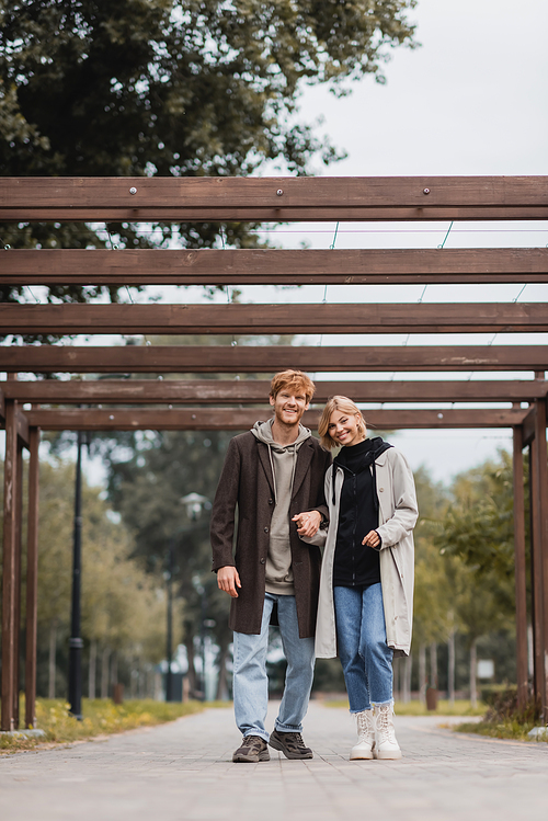 full length of joyful couple in autumnal coats holding hands while walking under multiple arch in park