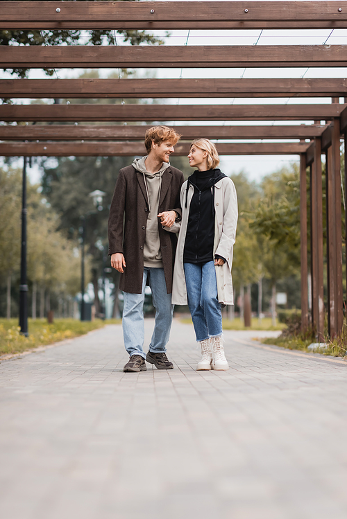 full length of happy couple in autumnal coats holding hands while walking under multiple arch in park