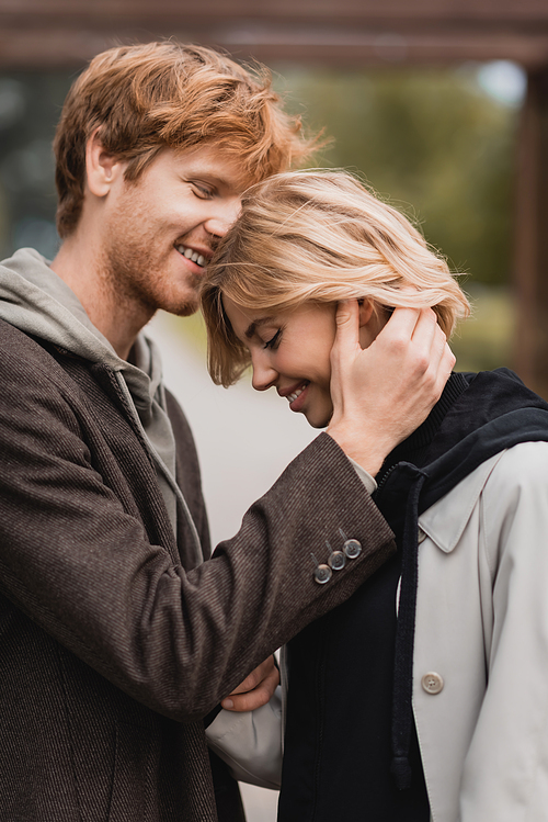 happy young man in autumnal coat hugging blonde girlfriend with closed eyes