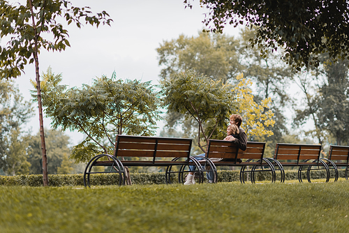 cheerful young man and woman hugging and sitting on wooden bench in green park