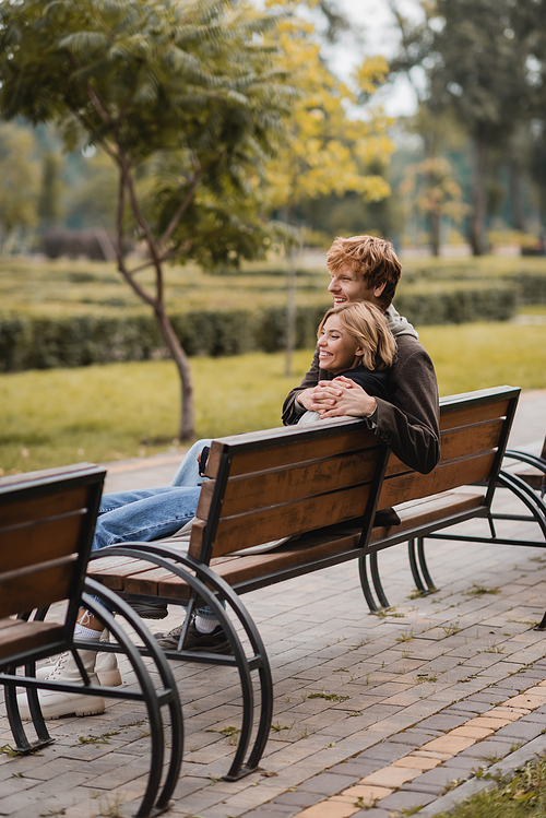 cheerful young man hugging woman and sitting on wooden bench in park