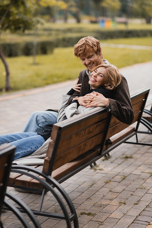 cheerful young man hugging positive woman and sitting on wooden bench in park