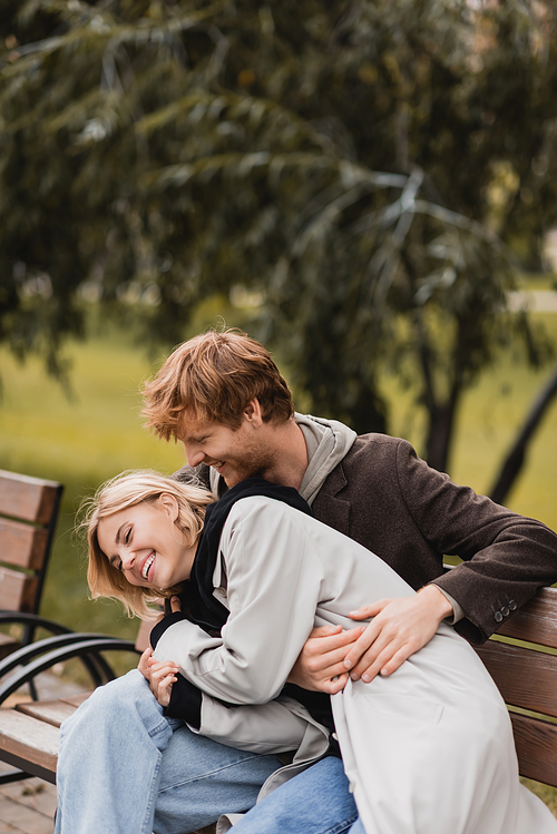 redhead man laughing while hugging happy girlfriend while sitting on bench