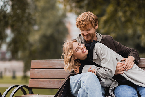 redhead man smiling while hugging cheerful girlfriend while sitting on bench