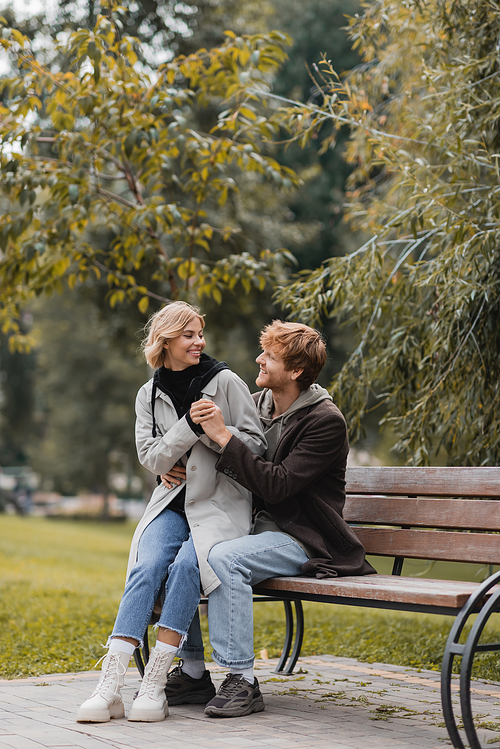 redhead man smiling while hugging positive girlfriend while sitting on bench