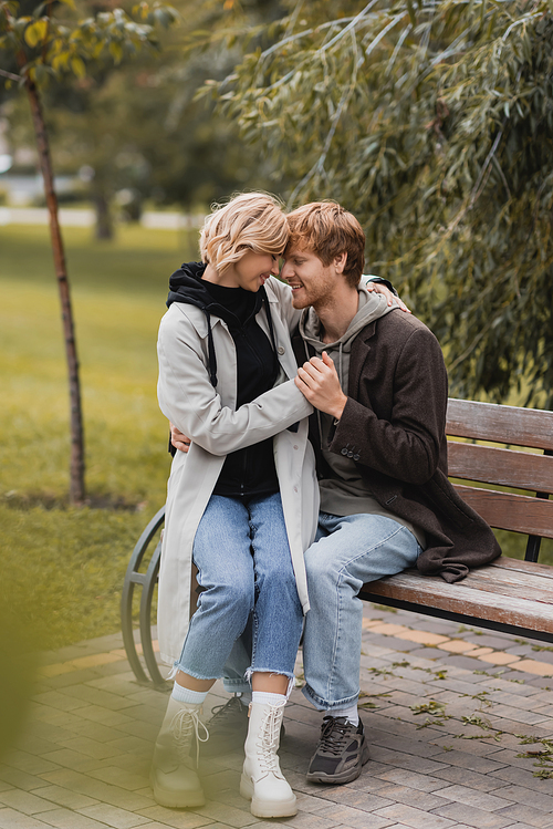 redhead man smiling while holding hands with positive girlfriend while sitting on bench