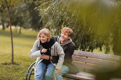 redhead man smiling while tickling positive girlfriend while sitting on bench