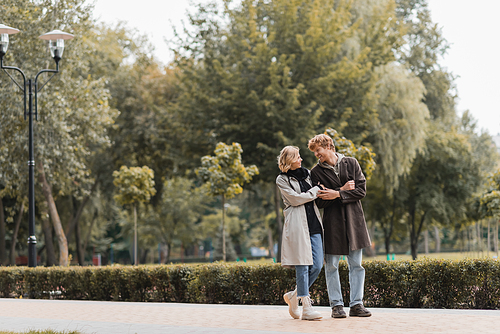 full length of redhead man and blonde woman in coat smiling while looking at each other in park