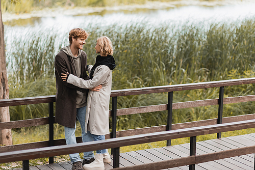 full length of redhead man and blonde woman in coat smiling while hugging on bridge near lake in park