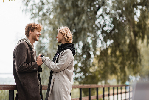 redhead man and blonde woman in coat smiling while holding hands near bridge in park