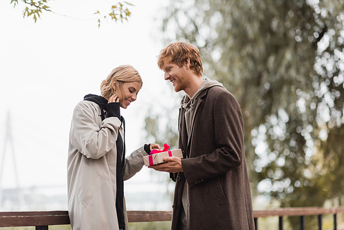 happy redhead man holding wrapped present near cheerful girlfriend in park