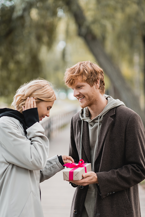 happy redhead man holding wrapped present near pleased girlfriend in park