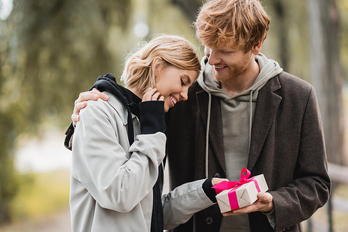 happy redhead man holding wrapped gift box near pleased girlfriend in park