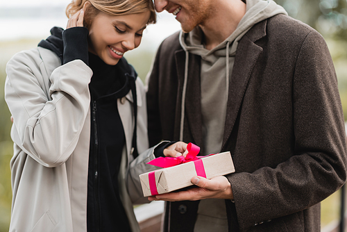 happy man holding wrapped gift box with pink ribbon near pleased girlfriend in park