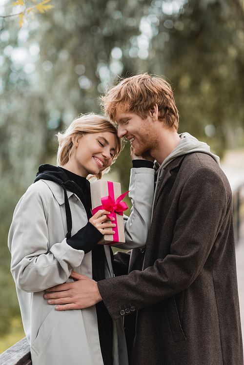 happy woman holding wrapped gift box with pink ribbon near pleased boyfriend in park