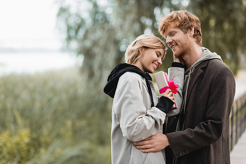 happy woman holding wrapped gift box with pink ribbon near cheerful boyfriend in park