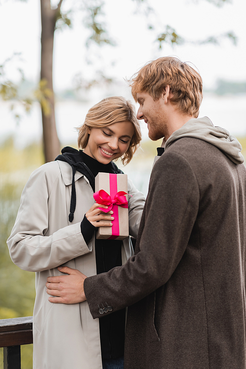 happy woman holding wrapped gift box with pink ribbon near joyful boyfriend in park