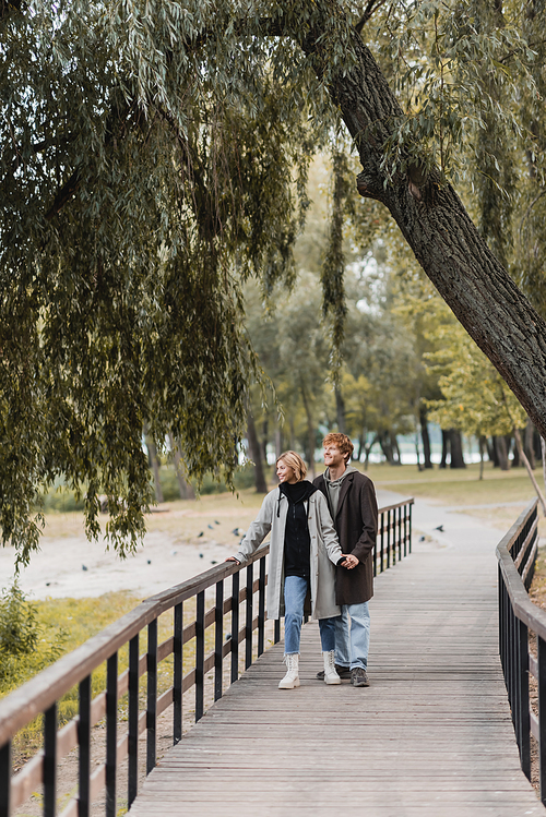 full length of redhead man and blonde woman in coat smiling while holding hands on bridge near pond in park