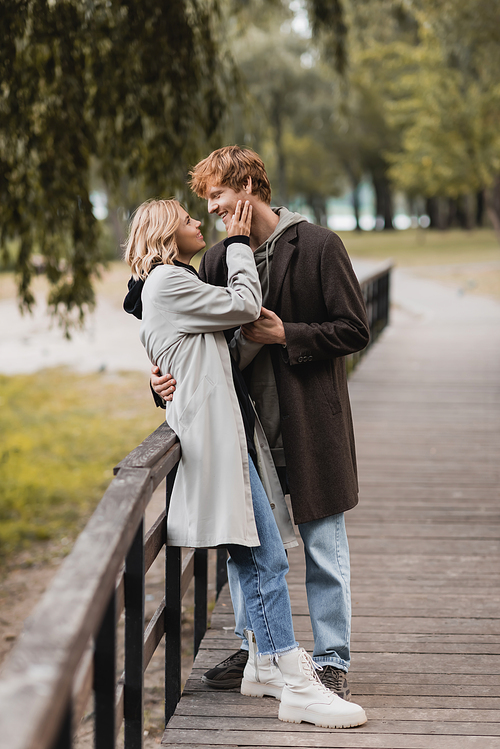 full length of redhead man and blonde woman in coat hugging while having date in park