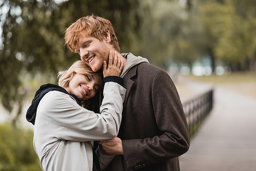 pleased redhead man and blonde woman in coat smiling while having date in park