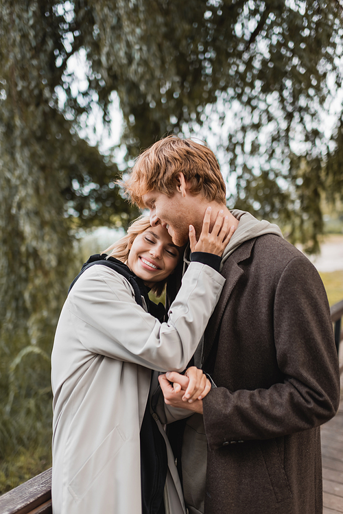 cheerful redhead man and pleased woman in coat smiling while having date in park