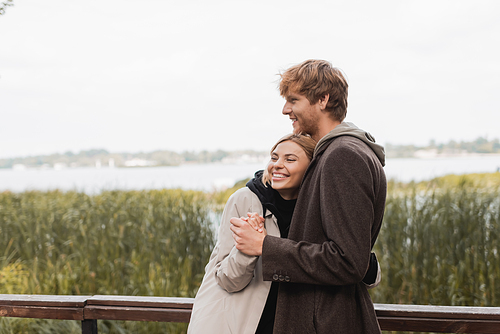 redhead man hugging and holding hand of cheerful blonde woman during date in park