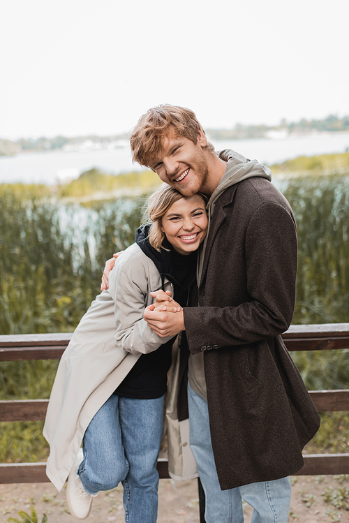 redhead man hugging and holding hand of cheerful blonde woman during date on bridge