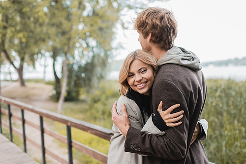 cheerful young woman smiling and hugging redhead man during date on bridge near pond